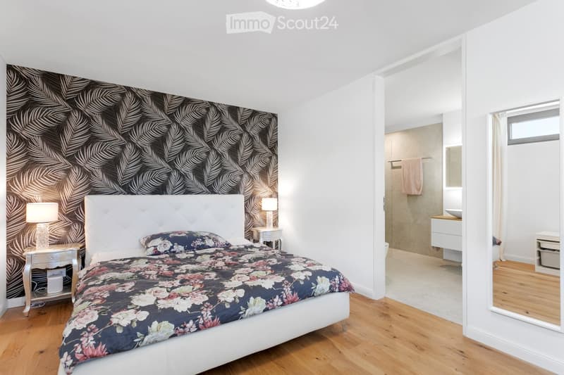 Modern bedroom with a white upholstered headboard, floral duvet, nightstands, and a lamp. Adjacent bathroom with a sink and mirror.