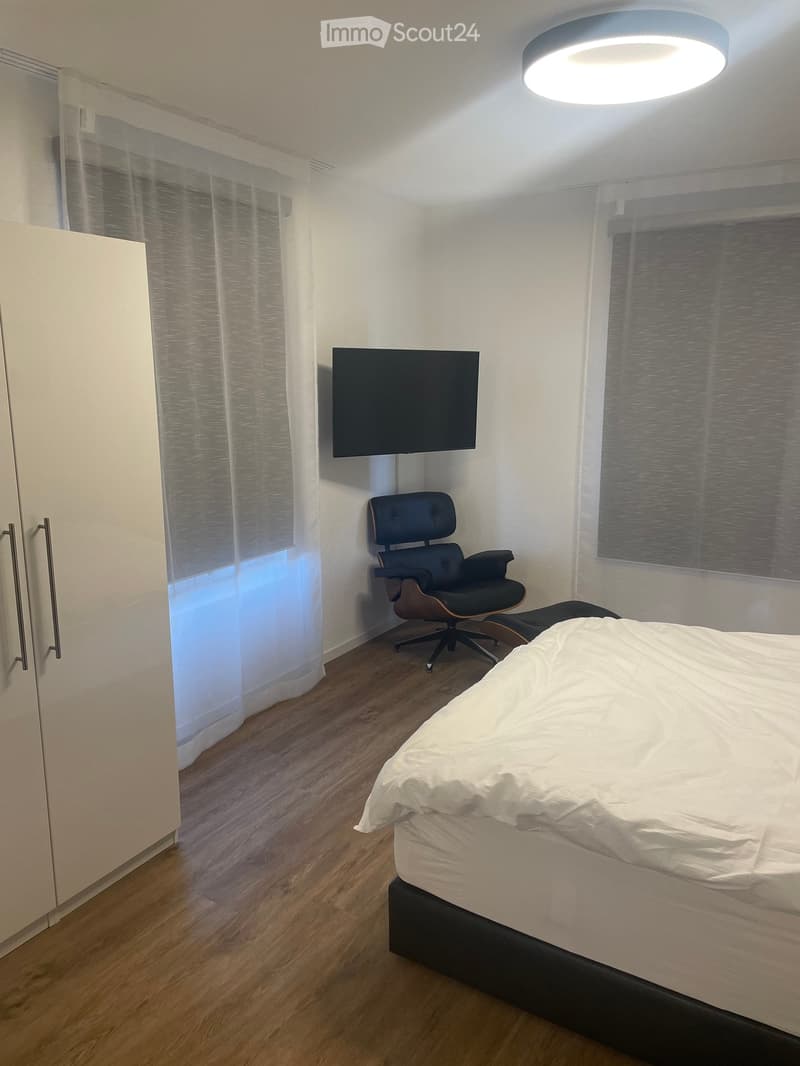 Modern bedroom with a wooden floor, white walls, a large bed with white bedding, a sleek black chair, and a mounted television on the wall.
