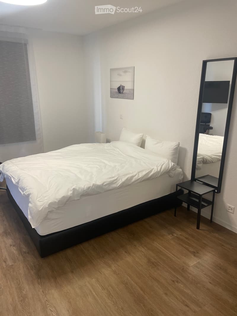 A modern bedroom with a neatly made bed, a large mirror, a nightstand, and a picture frame on the wall.