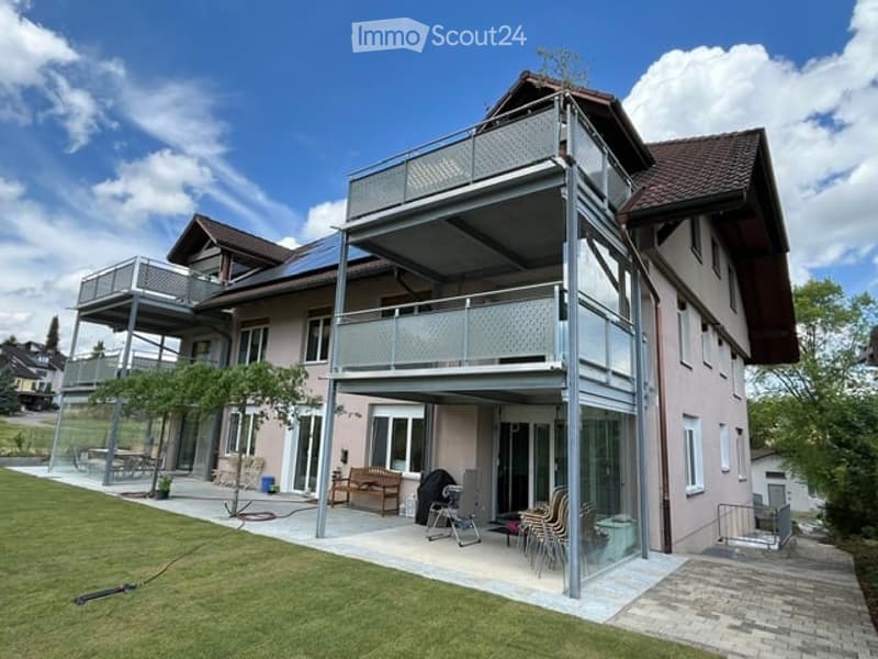 Modern two-story house with a large balcony and patio area, featuring outdoor furniture and a well-maintained lawn.