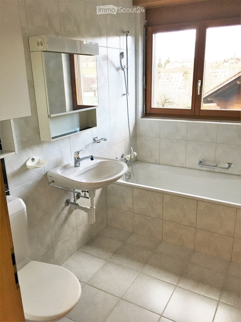 Modern bathroom with white tiles, a sink, bathtub, toilet, and a mirror cabinet. Features a large window with wooden frame and a view of the outside.