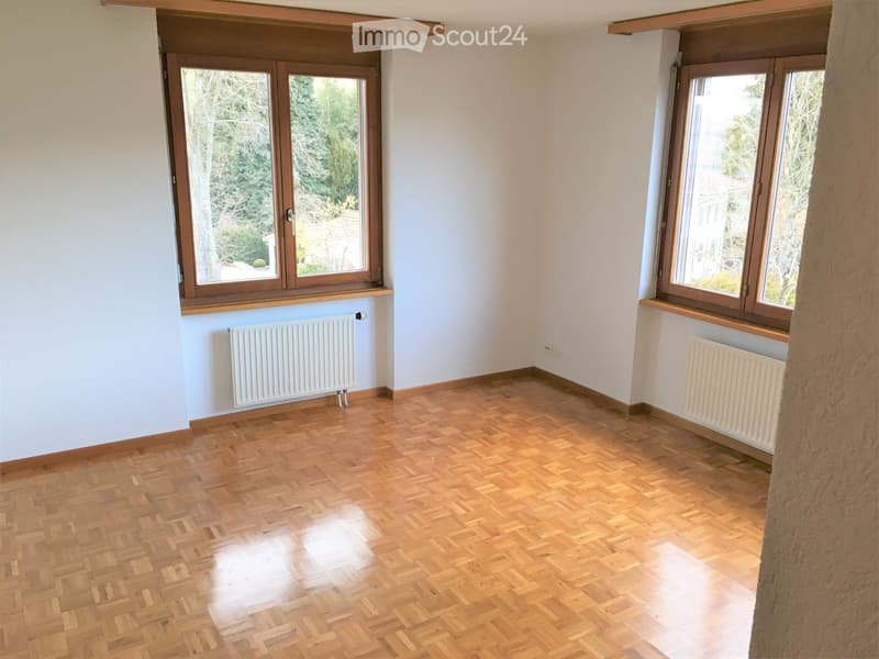 Empty room with wooden parquet flooring, white walls, and large windows with wooden frames. Radiators are visible beneath the windows.