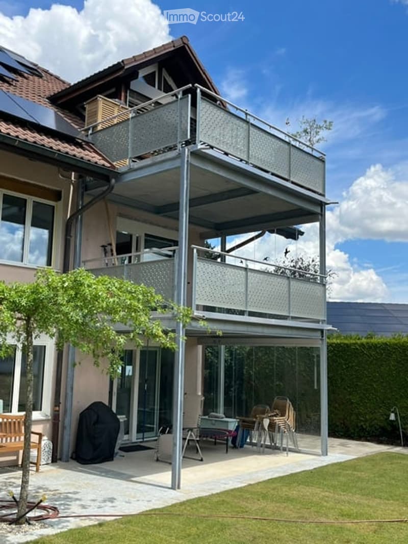 Modern outdoor area with a covered patio, metal railings, and a small seating area with chairs and a table. There is a wooden bench, a tree, and a covered balcony above.