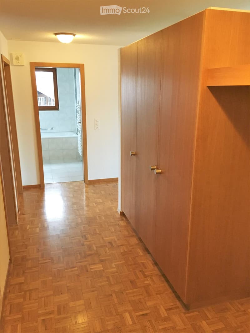 Empty hallway with wooden herringbone flooring, white walls, and a closed wooden door on the right. An open door on the left leads to a tiled bathroom with a bathtub and shower area.