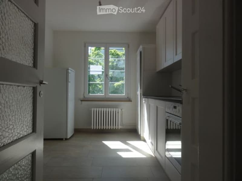 Empty kitchen with white cabinets, a window, a radiator, and a refrigerator. No furniture or appliances are present.