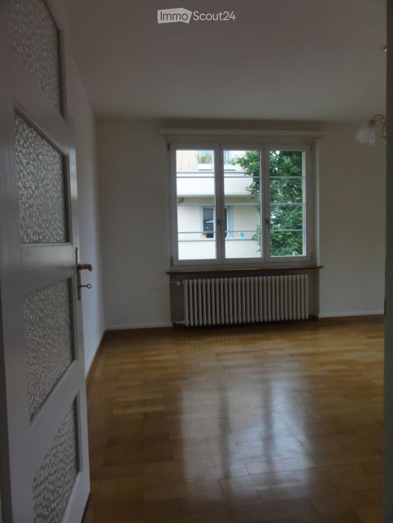 Empty room with wooden flooring, white walls, large window with radiator below, and a partially visible door with patterned panels.