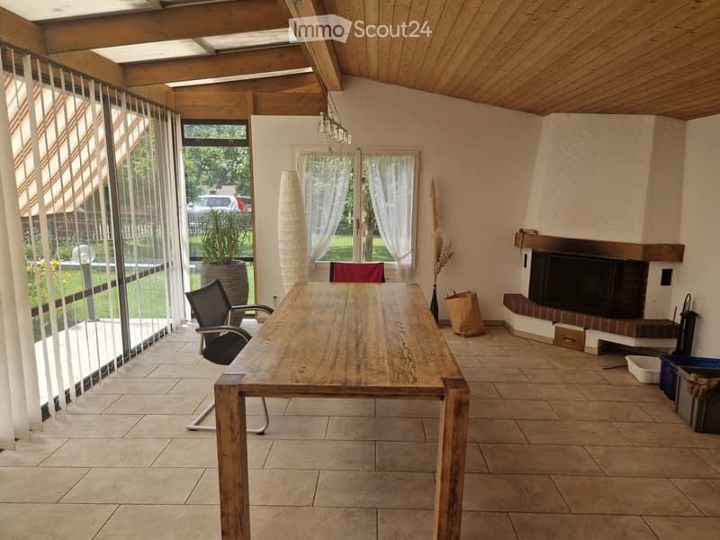 Bright, open room with wooden ceiling beams, large windows, and a fireplace. Features a long wooden table, office chair, and decorative elements like a vase and plant.