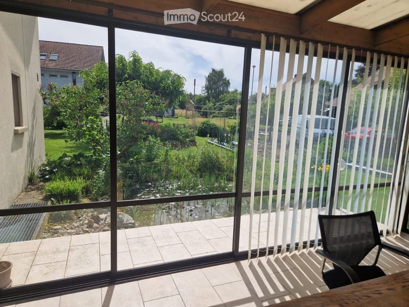 Balcony with large glass windows, tiled floor, and a view of a garden with a pond, trees, and a house in the background.