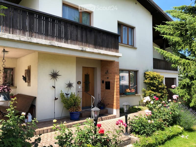 Two-story house with white exterior, wooden balcony, brick entrance, and well-maintained garden with flowers and shrubs.
