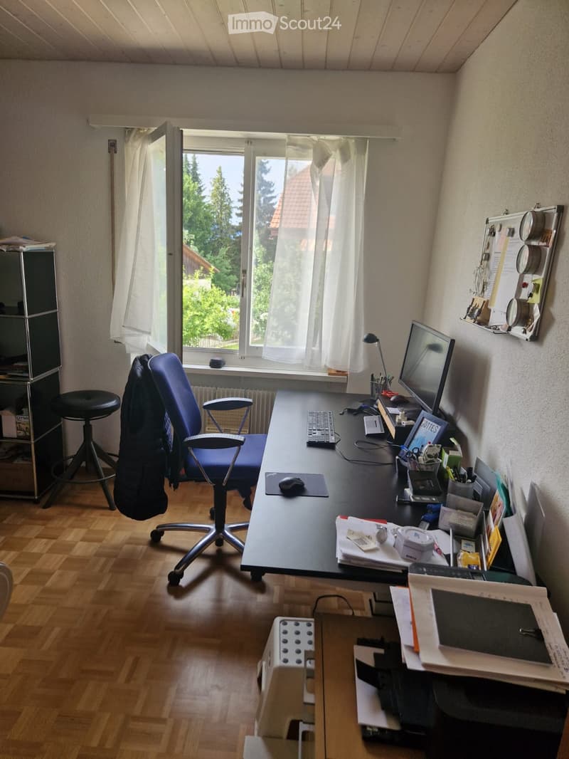 A well-lit office space with a large wooden desk, ergonomic chair, computer setup, and various office supplies. The room features wooden flooring and large windows with white curtains, offering a view of greenery outside.