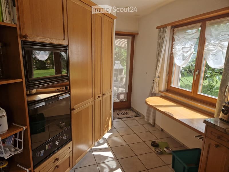 Bright kitchen with wooden cabinets, built-in oven, tiled floor, and large windows with white curtains. A small table and pet bowls are visible.