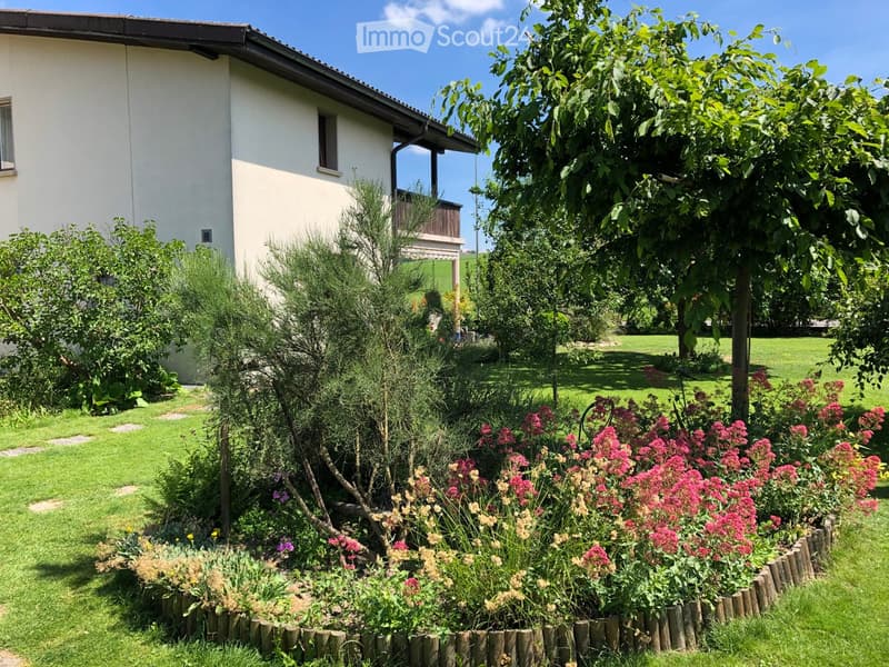 A well-maintained garden with a variety of flowering plants, shrubs, and trees surrounding a two-story house with a balcony and a visible window.