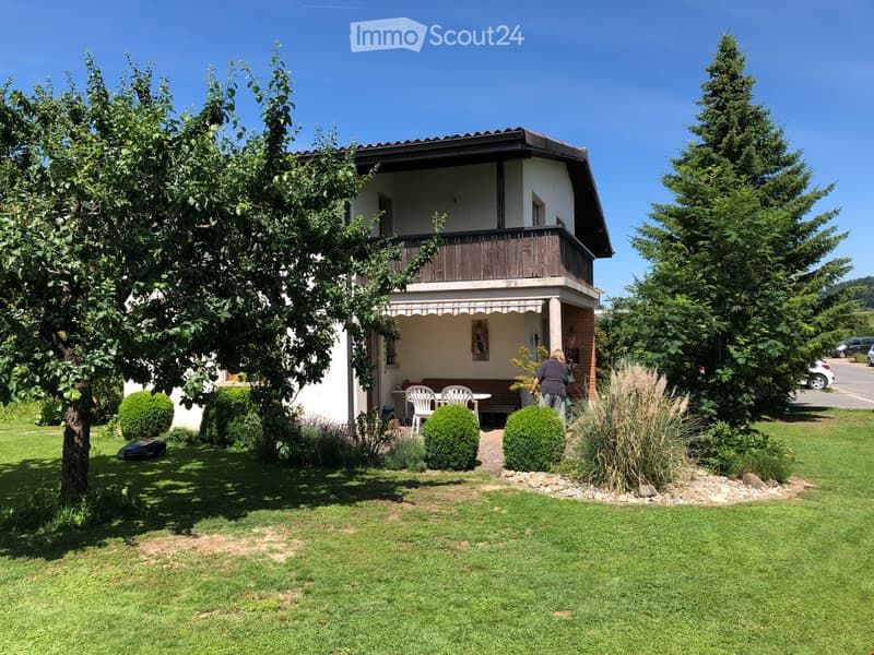 Two-story house with a balcony, surrounded by a well-maintained garden with a patio area, table, and chairs.