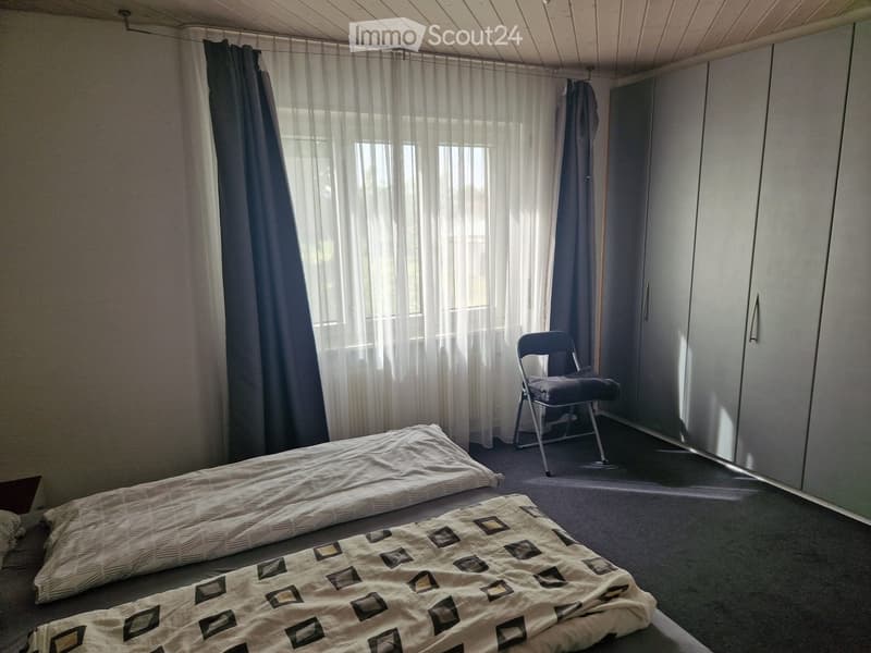 Small bedroom with two beds, a chair, and a wardrobe. The room has large windows with sheer curtains and a slanted ceiling.
