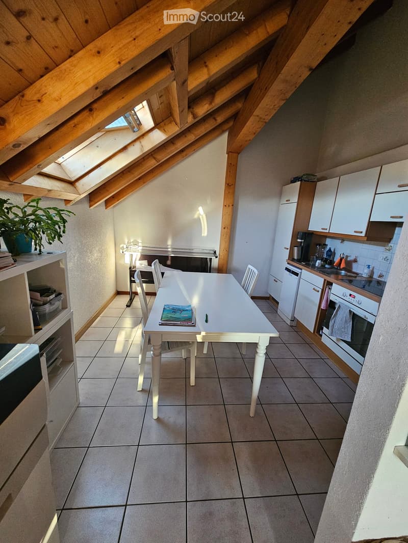 Bright kitchen with white cabinets, a dining table with chairs, and modern appliances including a refrigerator and oven. The room features exposed wooden beams and a skylight, with a small plant on a shelf.