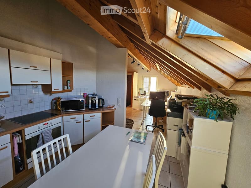 Open-plan kitchen and dining area with white cabinets, a dining table with chairs, and a workspace with a chair and printer. The kitchen features an oven, microwave, and coffee maker, with a skylight above.