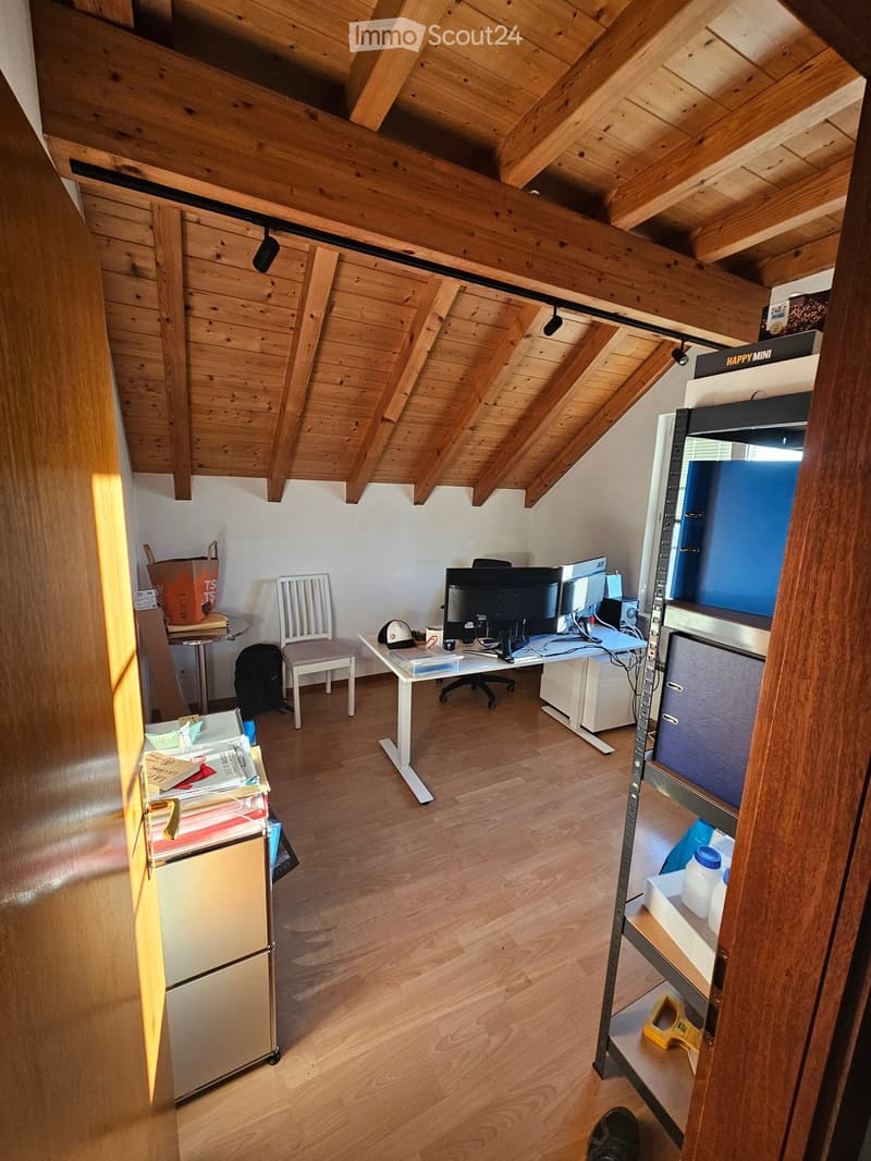 A furnished room with wooden beams on the ceiling, a desk with a laptop and screen, a chair, a small table with a cup, and a shelf with various items.