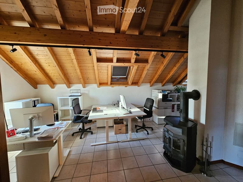 Office space with wooden ceiling beams, tiled floor, desk with computer, chairs, printer, and wood stove.