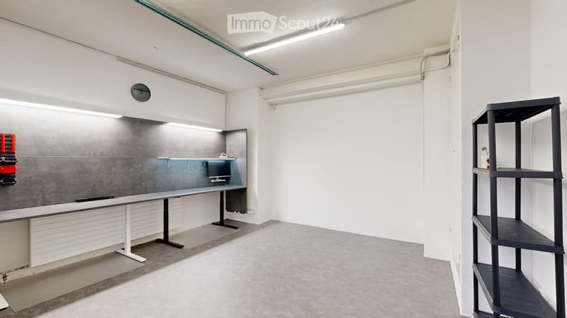 A modern, minimalist room with a grey wall, a white table and chair, a black shelf, a clock, and a screen on a grey counter.