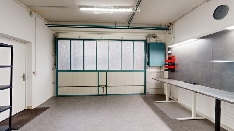 Empty room with a large garage door, white walls, grey floor, and a white table with a black surface. There is a shelf on the left side and a clock on the ceiling.