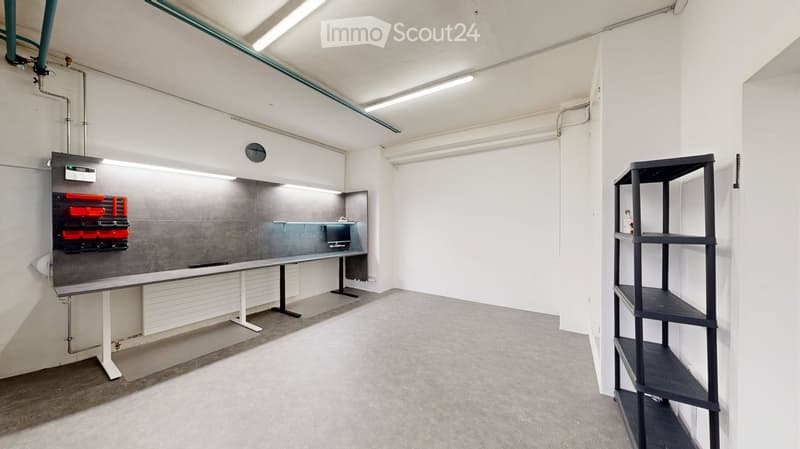 A clean, modern workshop or studio space with a large grey workbench, a black shelving unit, and a mounted screen.
