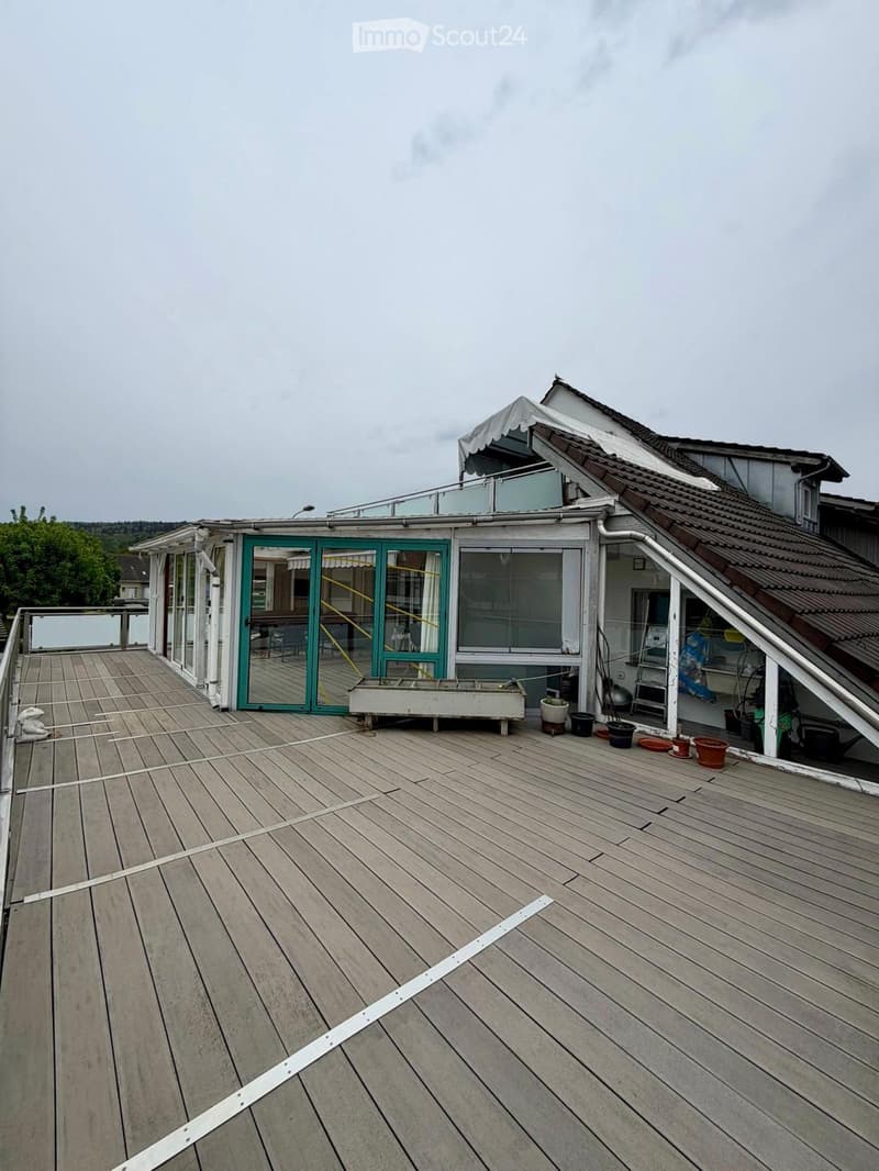 Wooden deck with a railing, large sliding glass doors leading to an indoor space, several potted plants, and a small covered area on the roof.