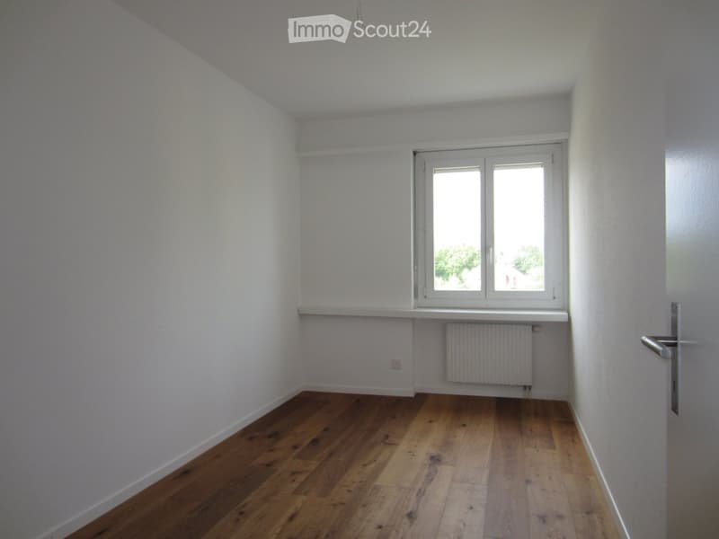 Empty room with wooden flooring, white walls, a window with white frame, a radiator, and a door with a silver handle.