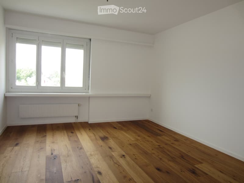 Empty room with wooden flooring, white walls, large windows, and a radiator under the windows.