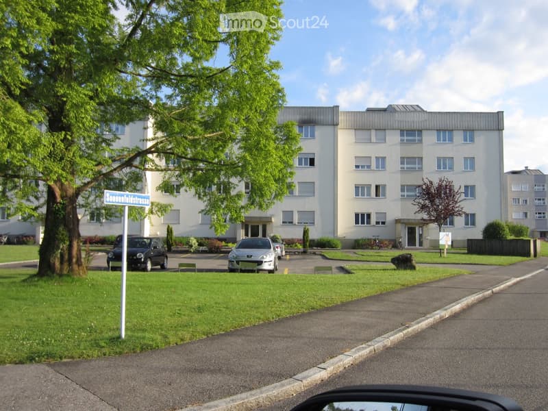 Multi-story residential building with white facade, multiple windows, and a parking area with several cars.