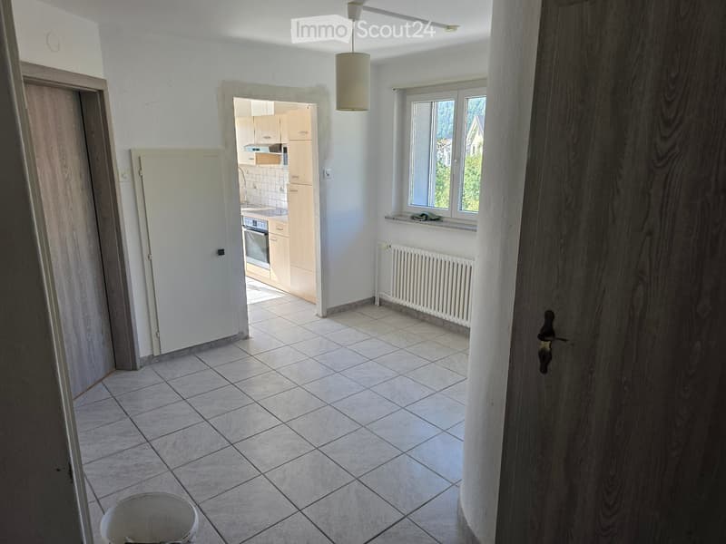 Empty room with tiled floor, white walls, and a view into a small kitchen area with cabinets and appliances.