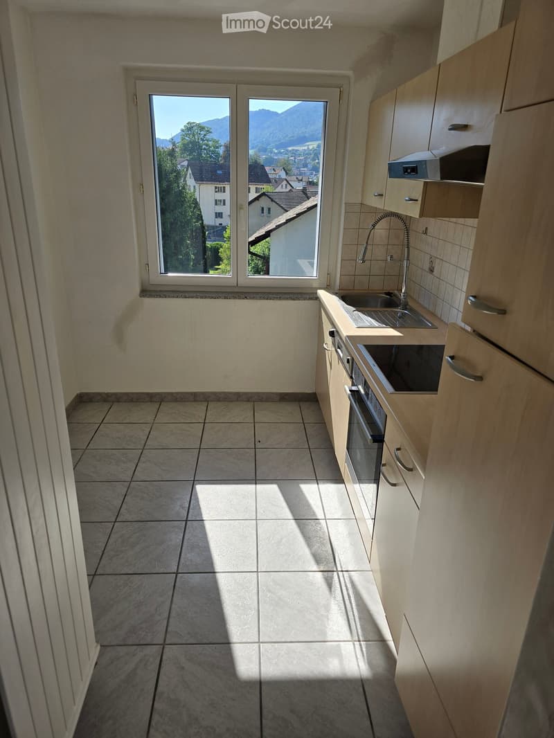 Modern kitchen with light wooden cabinets, tiled floor, and a window offering a scenic mountain view.