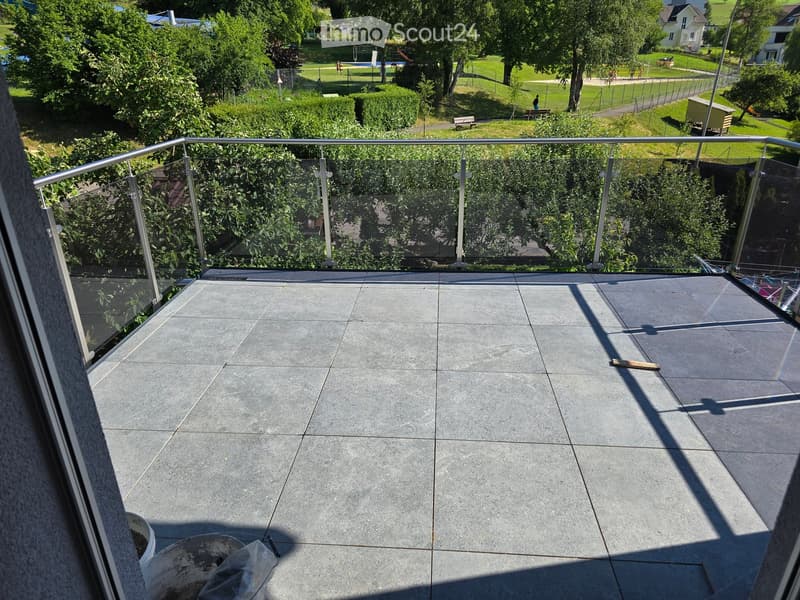 Balcony with grey tiled floor, glass railing, and view of a park with trees, benches, and a playground.
