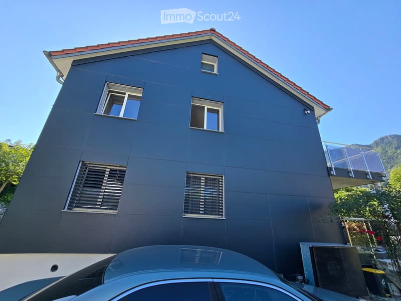 Modern two-story house with dark blue exterior, red-tiled roof, multiple windows, and a glass balcony on the upper level.