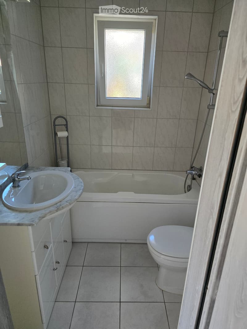 Modern bathroom with a white tiled floor and walls, featuring a sink with a marble countertop, a bathtub with a handheld shower, a toilet, and a window with frosted glass.
