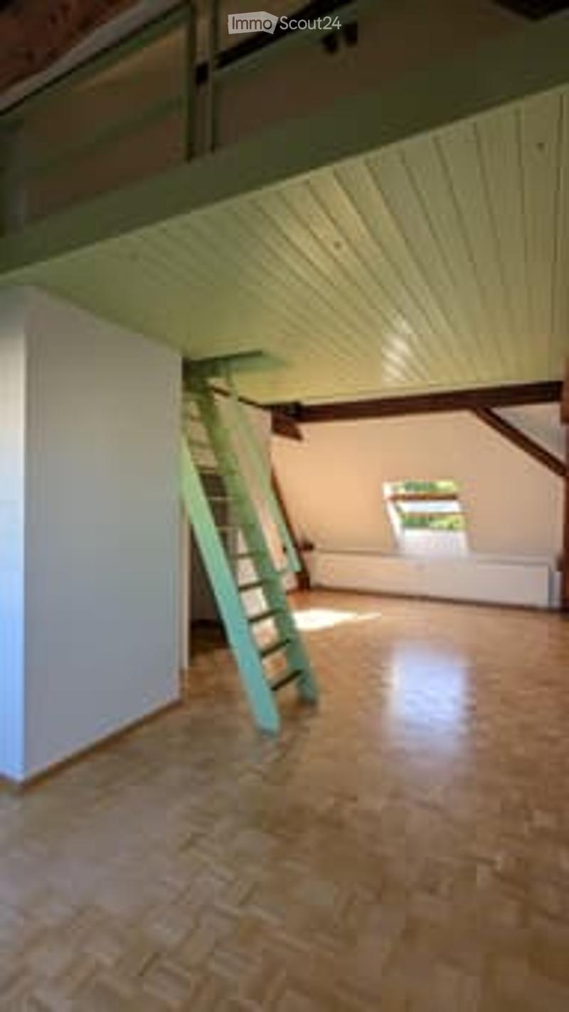 Empty attic room with wooden floor, white walls, exposed wooden beams, and a small window. Ladder leads to the upper level.