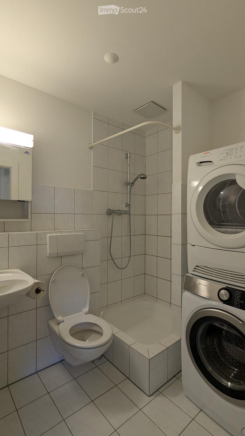 Modern bathroom with a toilet, sink, bathtub, shower, and a private washing machine. Light-colored tiles and a clean, organized layout.