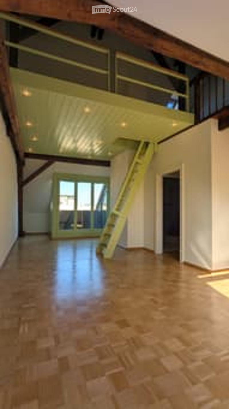 Bright, open loft space with exposed wooden beams, polished wooden floor, large windows, and a small staircase leading to an upper level.