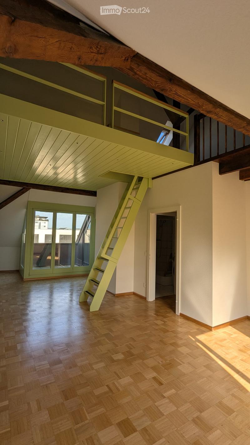 Bright, open loft space with wooden floors, exposed wooden beams, a green staircase leading to an upper loft area, and large windows letting in natural light.