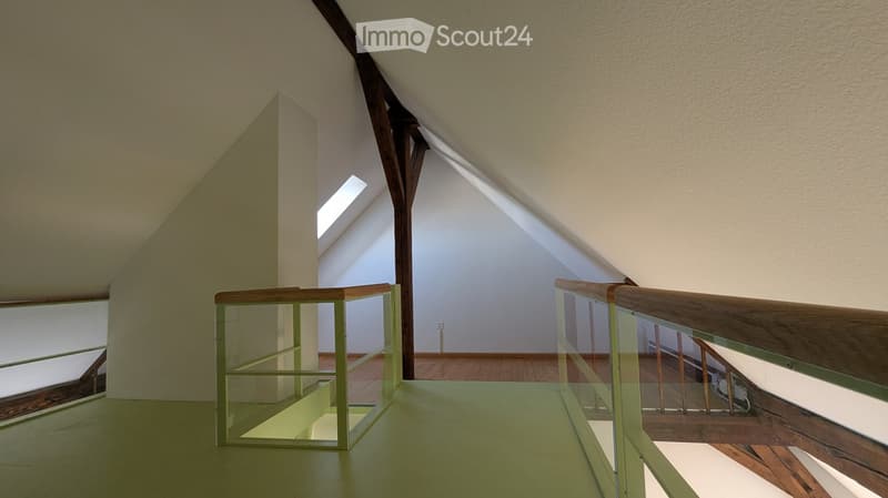 Modern hallway with glass railings, wooden beams, and exposed wooden floorboards. Natural light streams in from a skylight above.