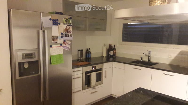 Modern kitchen with white cabinets, stainless steel appliances, and a dark countertop. Features include a refrigerator with photos and notes, an oven, a sink with a faucet, and several bottles on the counter.