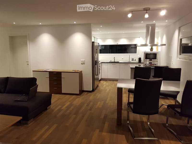 Modern open-plan living and dining area with a dark grey sofa, laptop on a coffee table, a glass dining table with four chairs, and a sleek kitchen featuring stainless steel appliances and white cabinets.