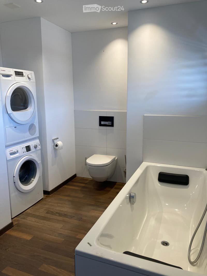 Modern bathroom with a bathtub, toilet, and Bosch washing machine. The room features white tiled walls, wooden flooring, and recessed lighting.