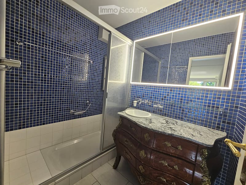 Elegant bathroom with blue mosaic tiles, marble countertop, ornate wooden vanity, and a large illuminated mirror above the sink.