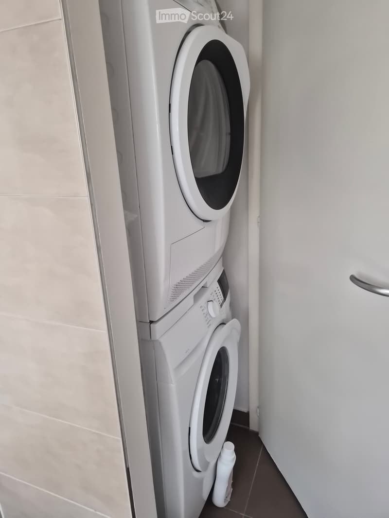 A compact bathroom with a stacked washing machine and dryer unit, light-colored tiles, and a small bottle placed on the floor next to the dryer.