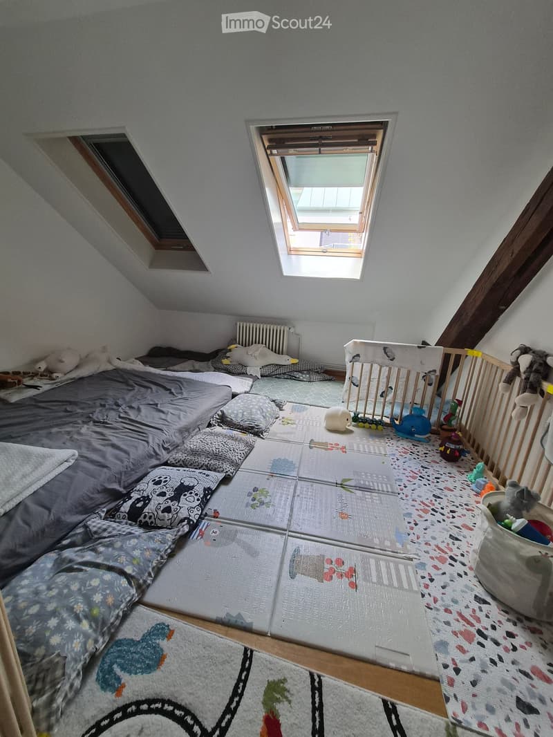 Cozy attic bedroom with a lofted bed, colorful pillows, a crib, and toys scattered around. The room features skylights and a patterned rug.
