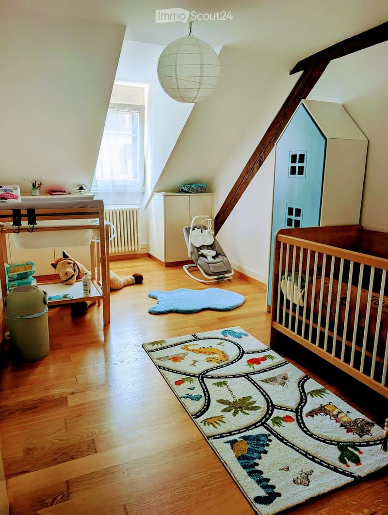 Cozy children's bedroom with wooden floor, crib, baby chair, toy house, and colorful rugs.