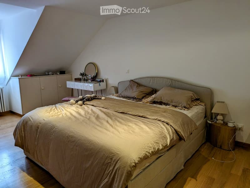 Cozy attic bedroom with wooden floors, a large bed with beige bedding, a nightstand with a lamp, a vanity table with a mirror, and a small cabinet.