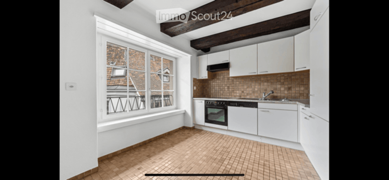 Bright, unfurnished kitchen with white cabinets, exposed wooden beams, and a large window offering a view of a tiled roof. The floor is made of warm, terracotta tiles.