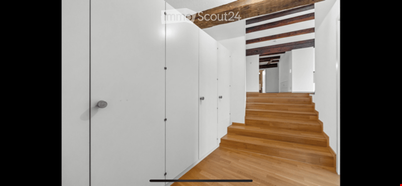 A modern hallway with white walls, wooden flooring, and a staircase leading upwards. The hallway features sleek white cabinetry on one side and exposed wooden beams on the ceiling.