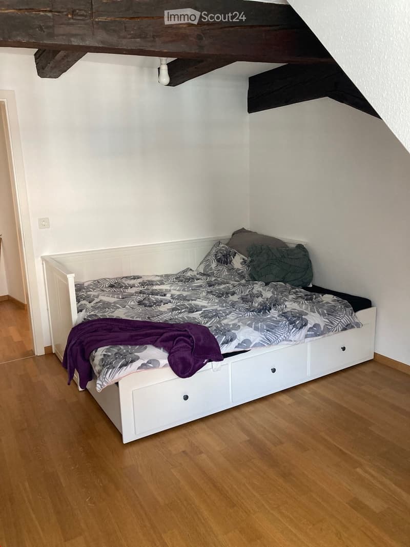 A small bedroom with a white bed featuring a grey and black patterned duvet, pillows, and a purple throw. The bed has built-in drawers and is placed on a wooden floor.